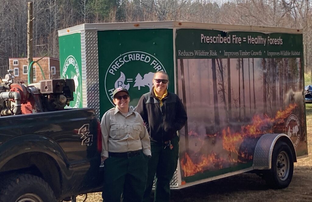 Two people stand in front of a Prescribed Fire trailer.